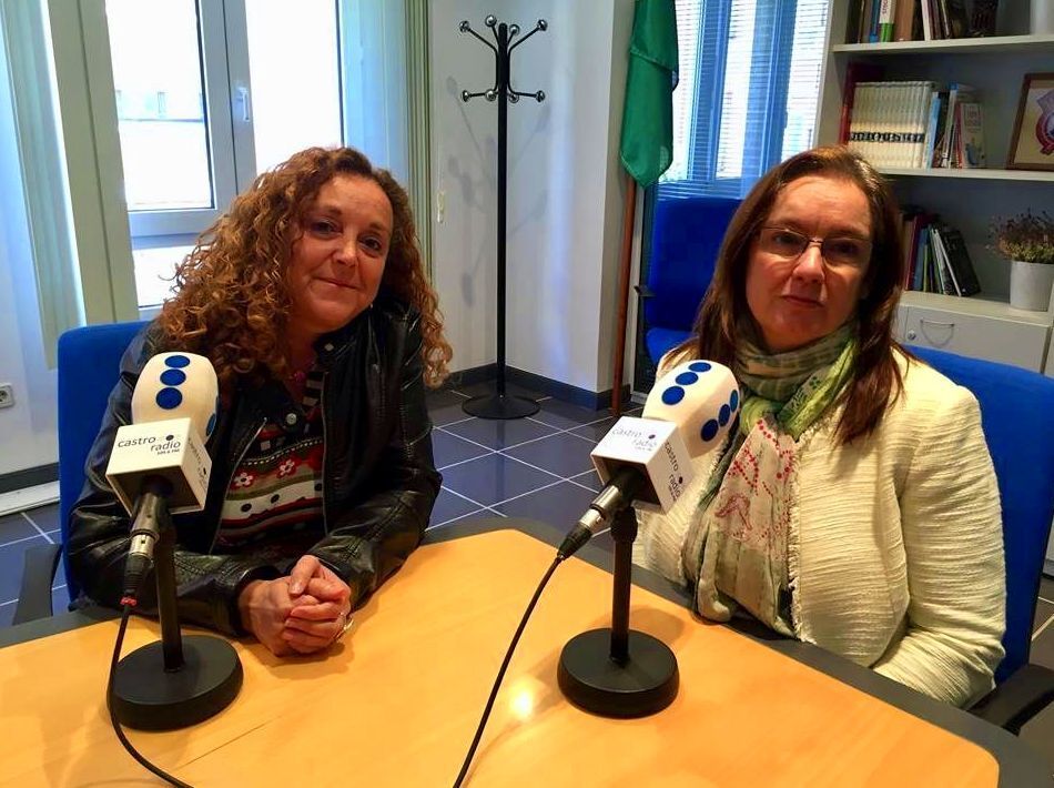 Dos mujeres sentadas en una mesa con micrófonos, probablemente para una entrevista de radio, en una oficina.