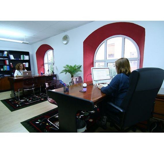 Dos mujeres trabajando en una oficina con escritorios. Paredes blancas, marcos de ventanas rojos y arqueados