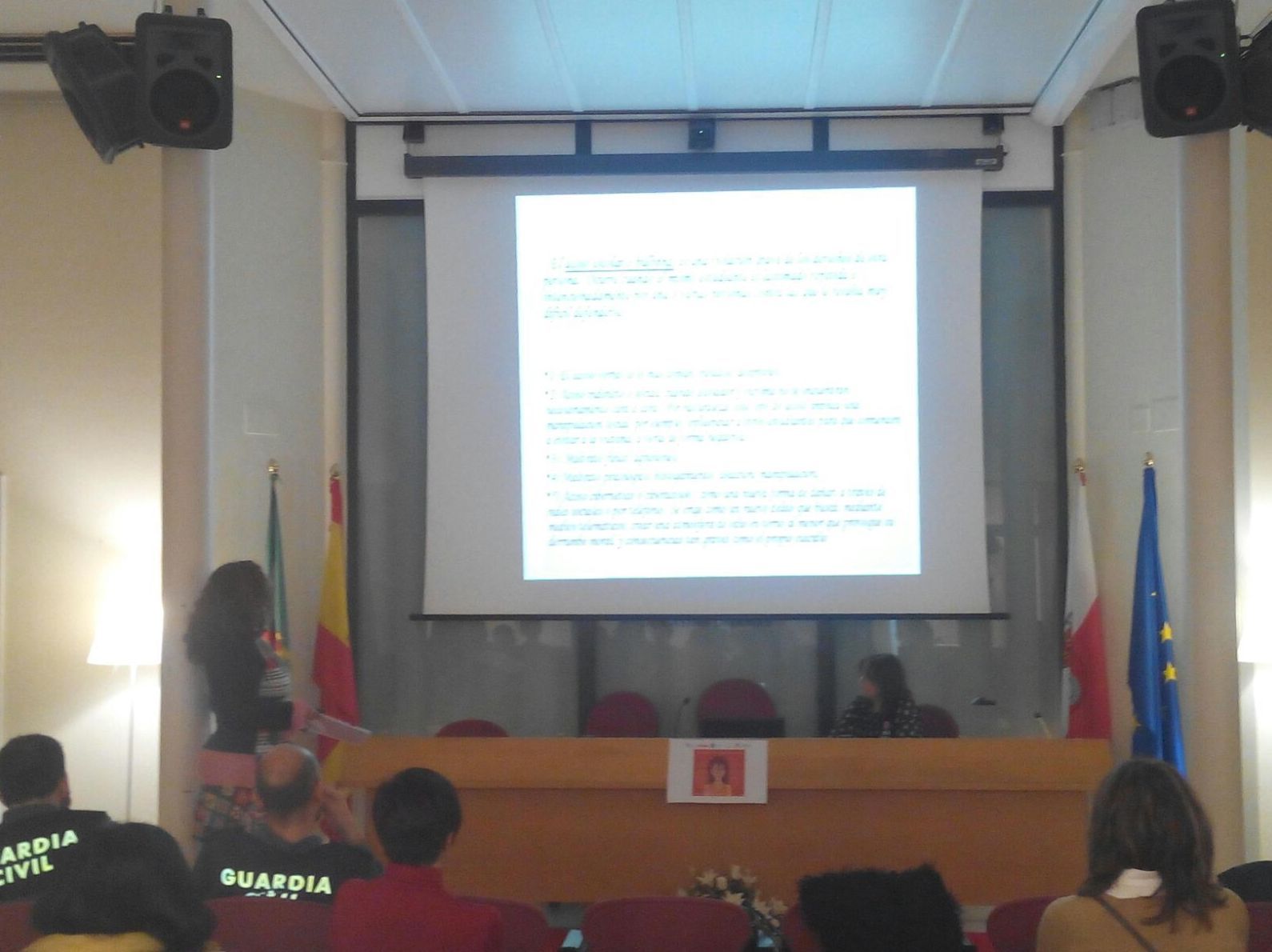 Una mujer presenta en una conferencia. Una pantalla muestra texto; se ven las banderas de España, Polonia y la UE. 