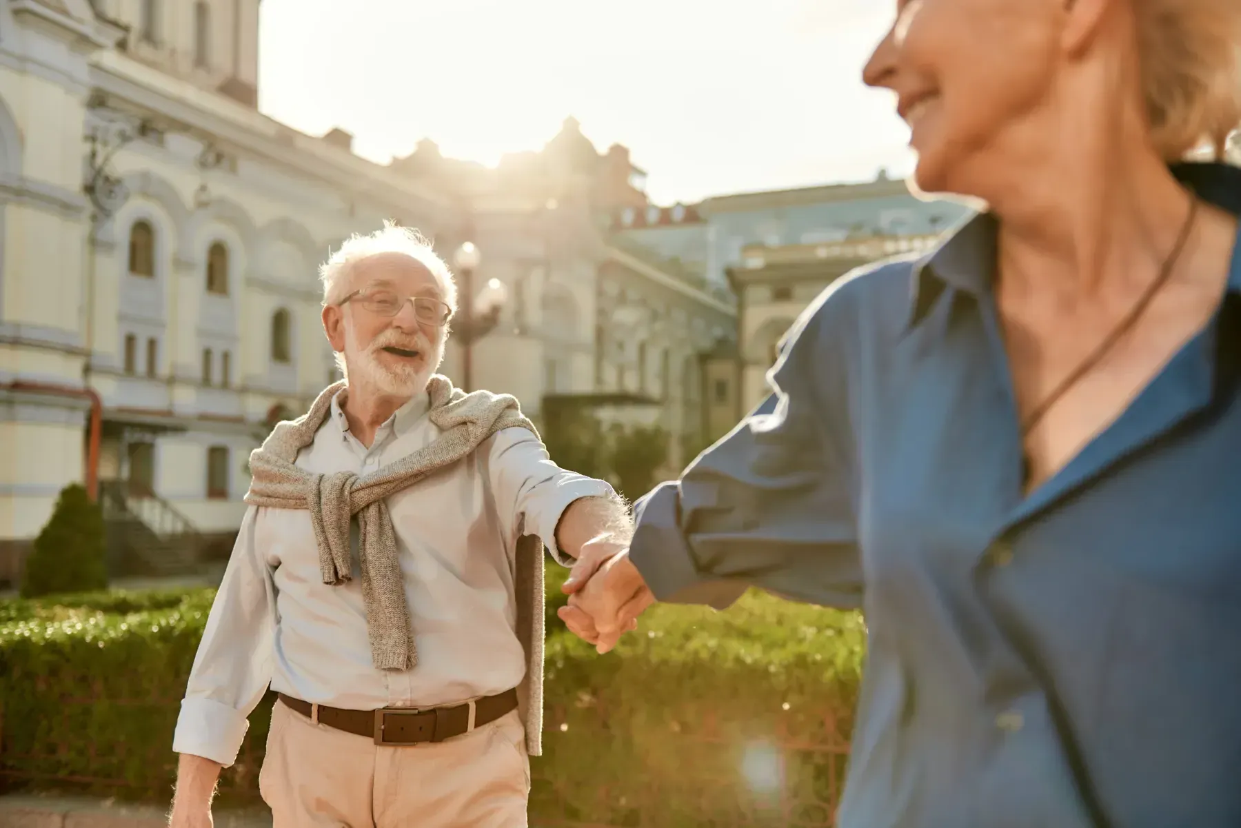Una pareja sonriente camina de la mano al aire libre, cerca de un edificio histórico de piedra, bajo la cálida luz del sol.