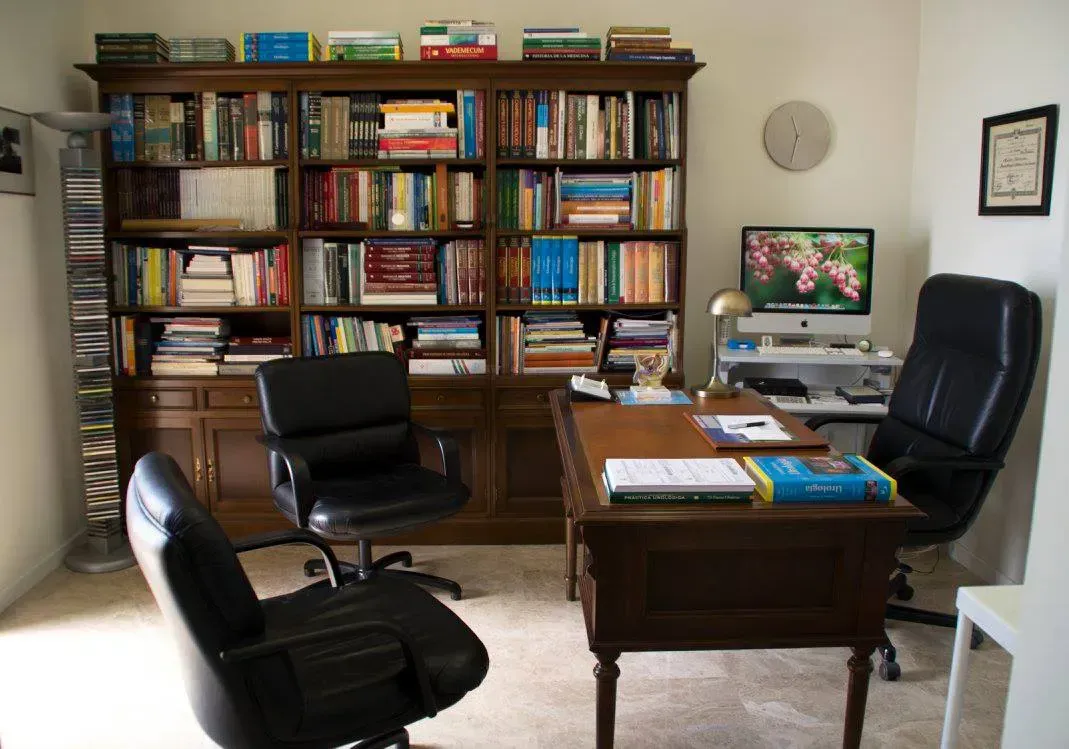 Un despacho en casa con un escritorio de madera, tres sillas negras, una gran estantería llena de libros