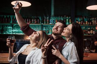 Tres amigos sonríen mientras se toman una selfie con una bebida en un bar con poca luz