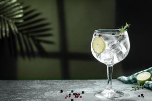 Un gin tonic en un vaso con una rodaja de lima y una ramita de romero, sobre una superficie de piedra con granos de pimienta.