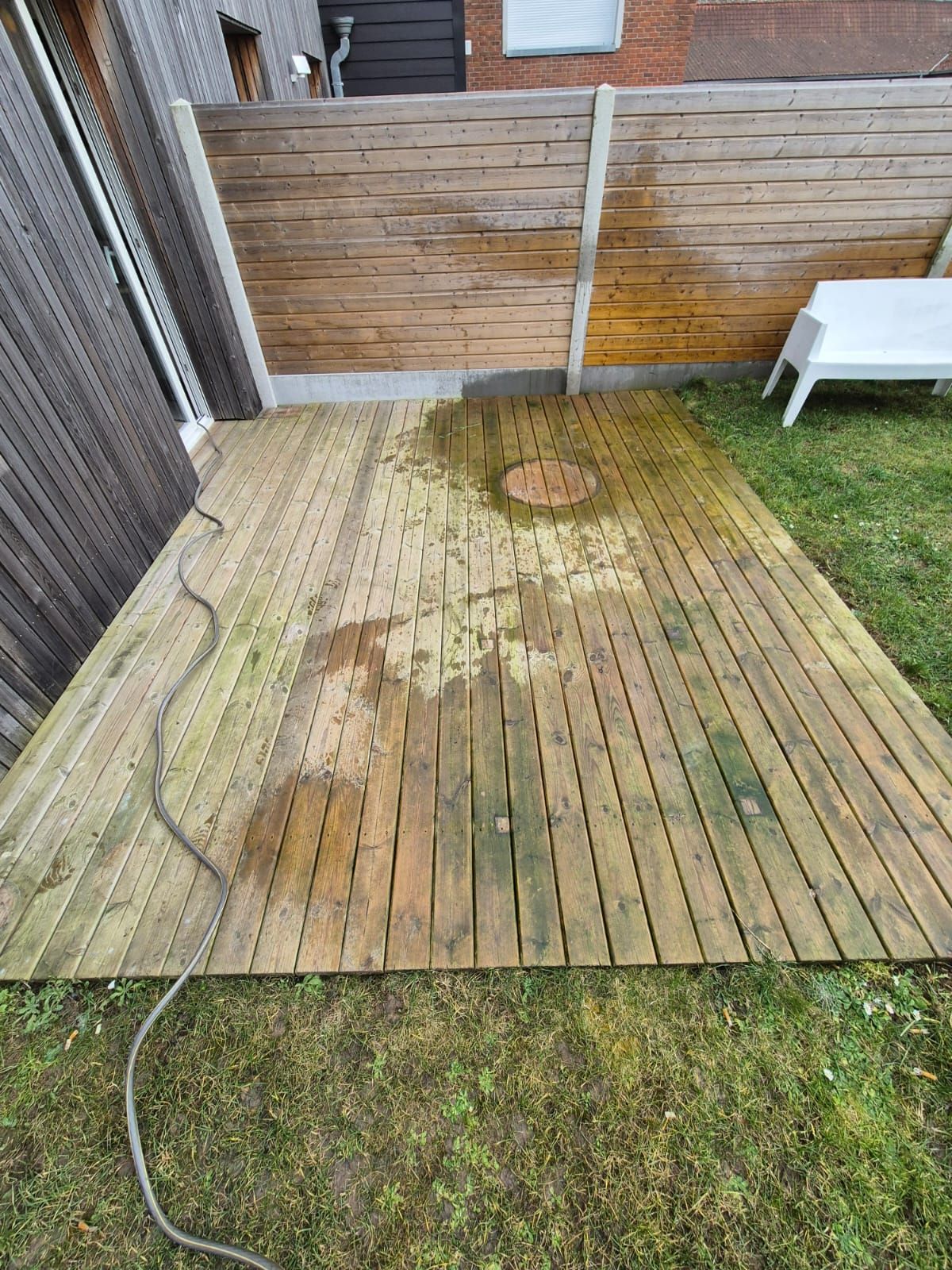 Terrasse en bois avec algues, tuyau d'arrosage sur gazon. Clôture et chaise blanche en arrière-plan.