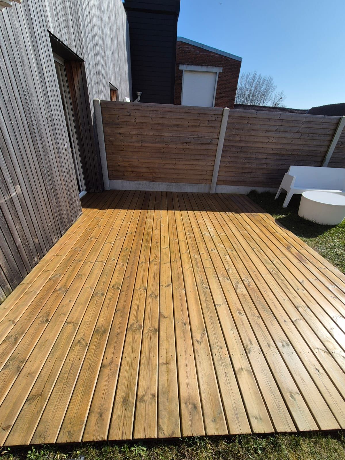Terrasse en bois dans un jardin, planches marron clair. Espace clôturé, ciel bleu, mobilier blanc.