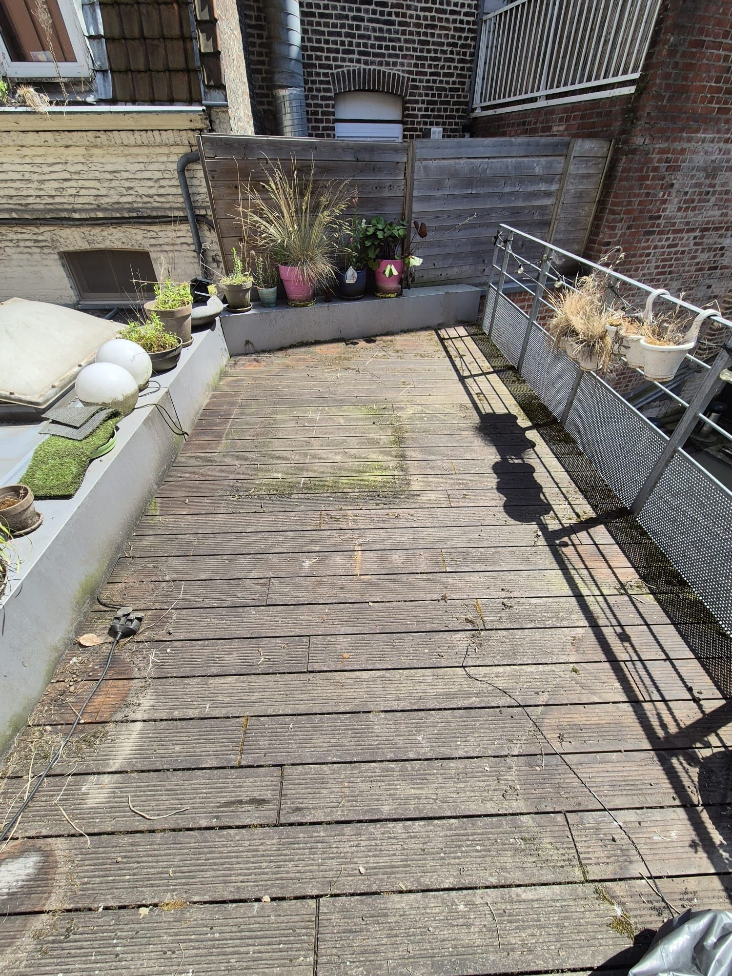 Terrasse sur le toit avec planches de bois patinées, plantes et balustrade en métal sous un ciel ensoleillé.