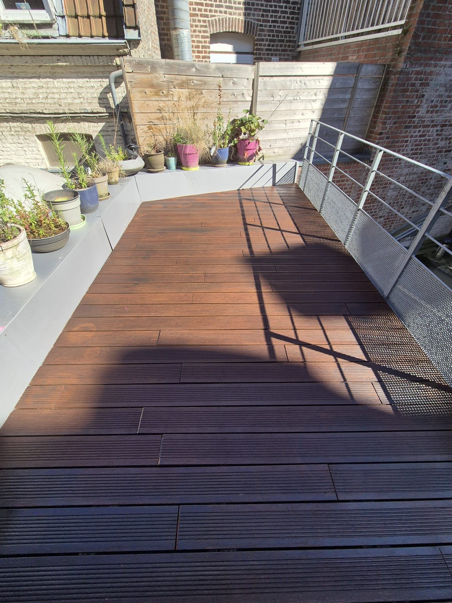 Terrasse en bois sur le toit avec des plantes en pot et une balustrade en métal par une journée ensoleillée.