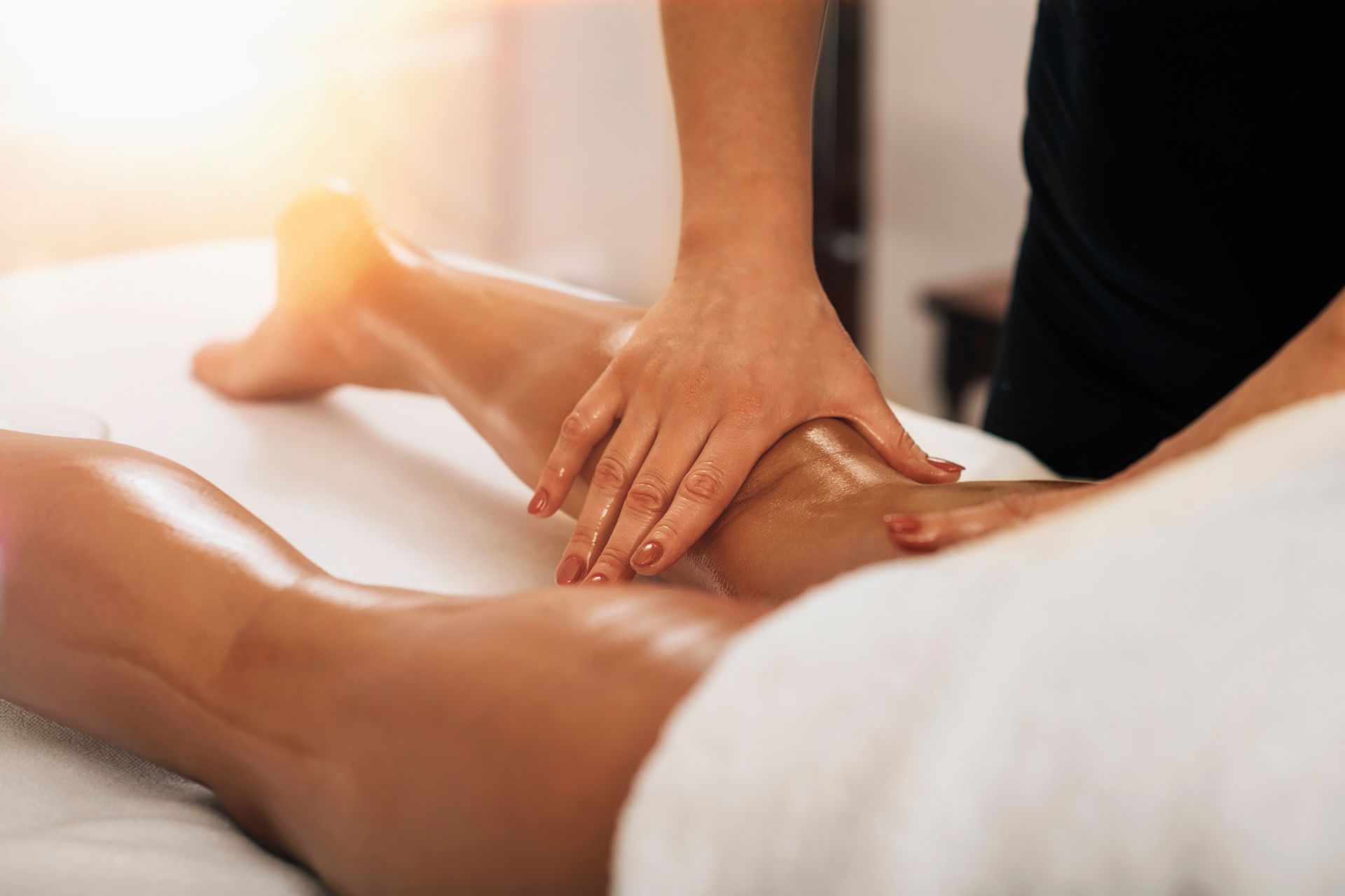 Massage des jambes : des mains massent une jambe sur une table de massage blanche, avec la lumière du soleil en arrière-plan.