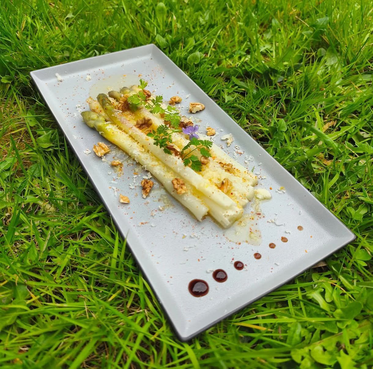 Une assiette rectangulaire d'asperges blanches, garnie de noix et d'herbes aromatiques, est posée sur une pelouse verte, agrémentée de points de vinaigre balsamique.
