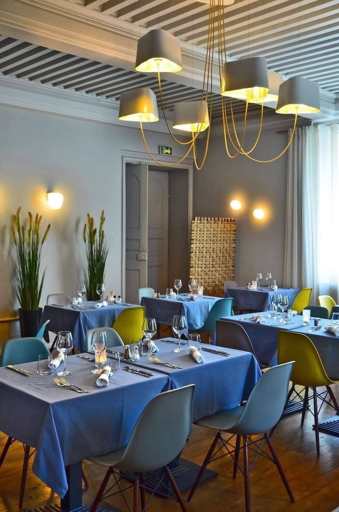 Une salle à manger lumineuse avec des tables nappées de bleu, des chaises colorées, un lustre moderne doré et de grandes plantes en pot.