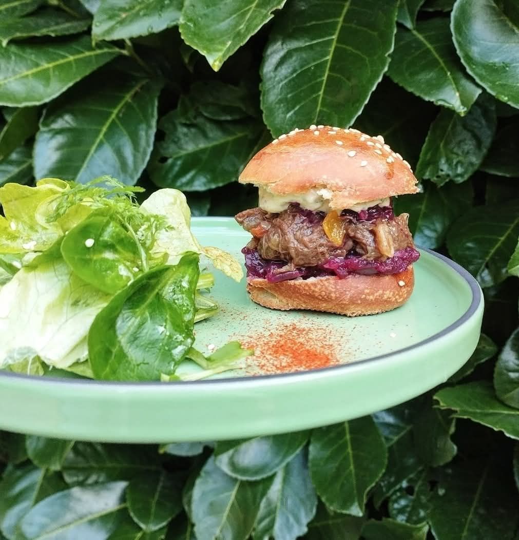 Un mini-burger garni de chou rouge et de sauce, accompagné d'une salade verte fraîche, le tout sur une assiette vert pâle devant un fond de feuillage.