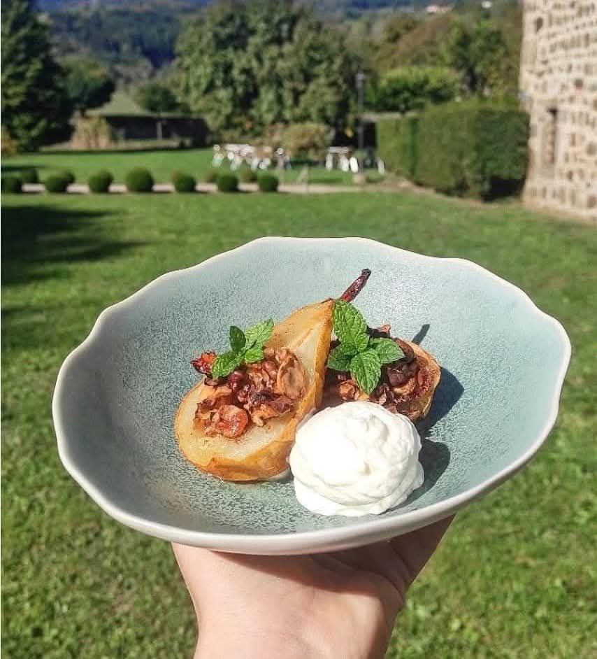 Une main tient un bol en céramique contenant deux moitiés de poires rôties, garnies de noix et de menthe, et servies avec une quenelle de crème.