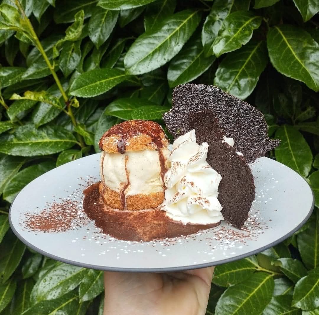 Une pâtisserie fourrée à la crème, nappée de sauce au chocolat, de crème fouettée et d'une gaufrette au chocolat noir, servie sur une assiette en extérieur.