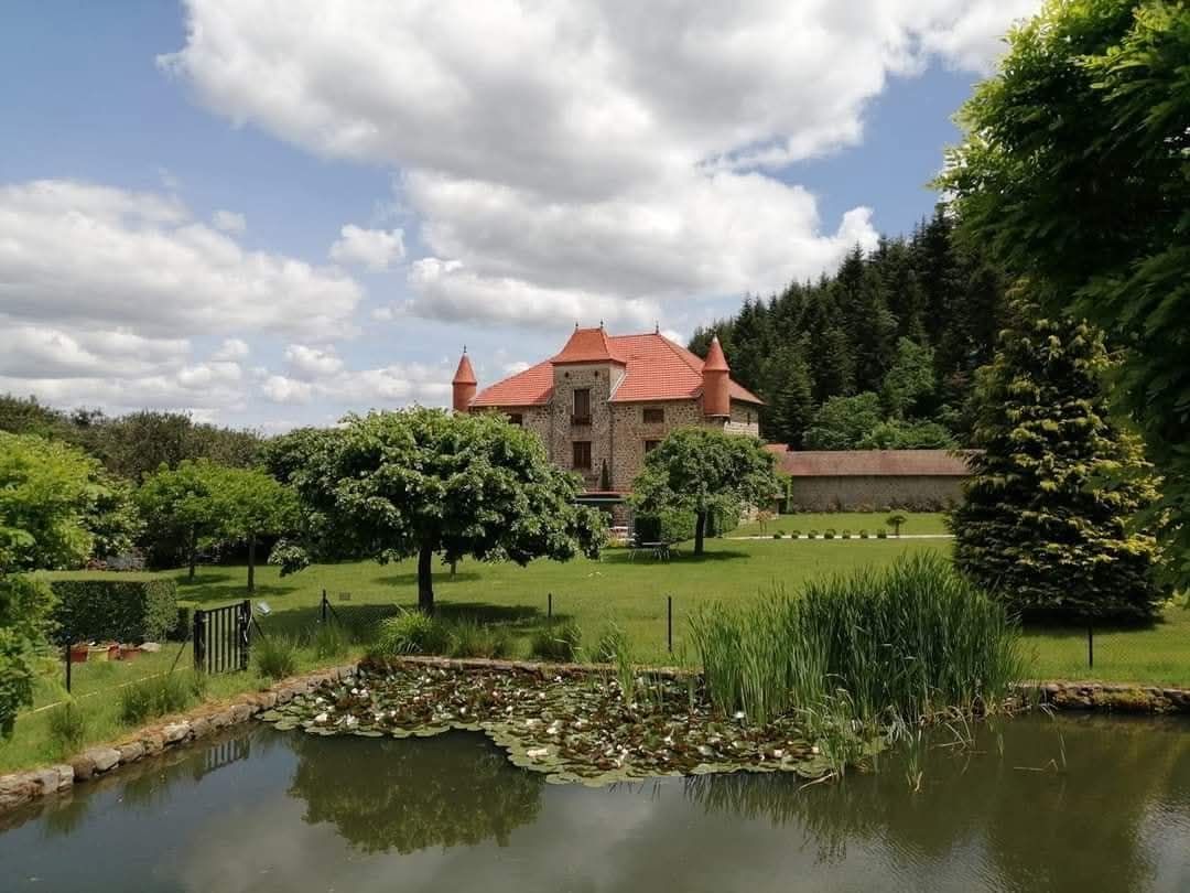 Un château de pierre au toit rouge se dresse derrière une pelouse verdoyante et un petit étang, entouré d'arbres sous un ciel nuageux.