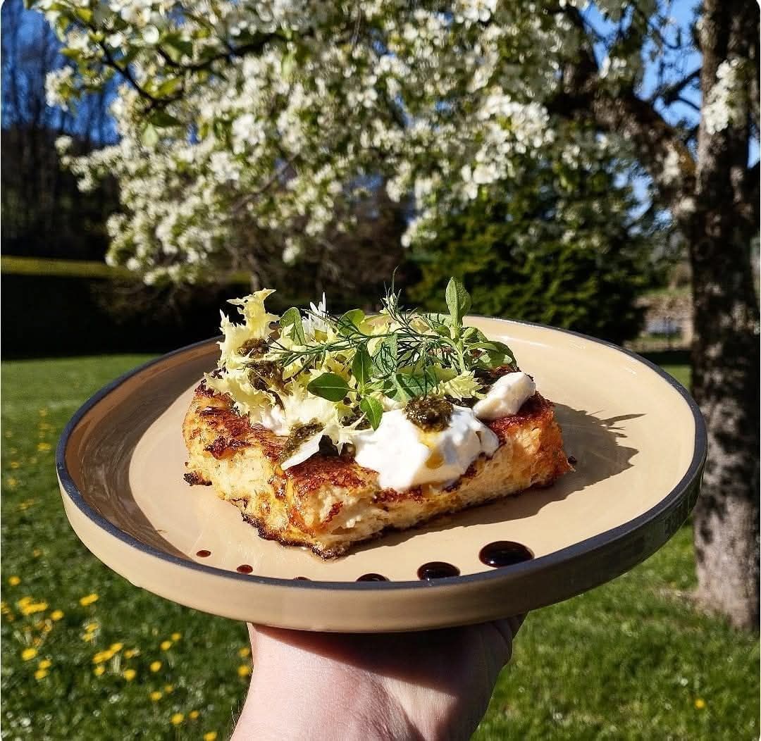 Une main tient une assiette de nourriture croustillante garnie d'herbes, de crème blanche et de glaçage balsamique, à l'extérieur, dans un jardin ensoleillé et fleuri.