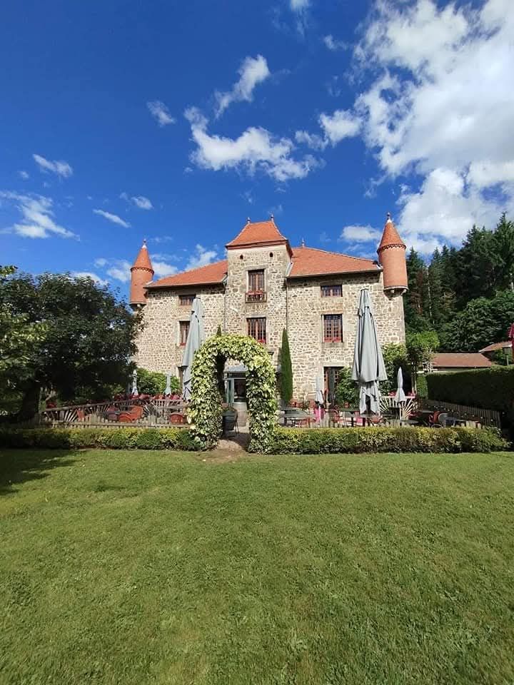 Un château en pierre au toit de tuiles rouges et à deux tourelles, encadré par une arche fleurie sur une pelouse verte sous un ciel bleu.