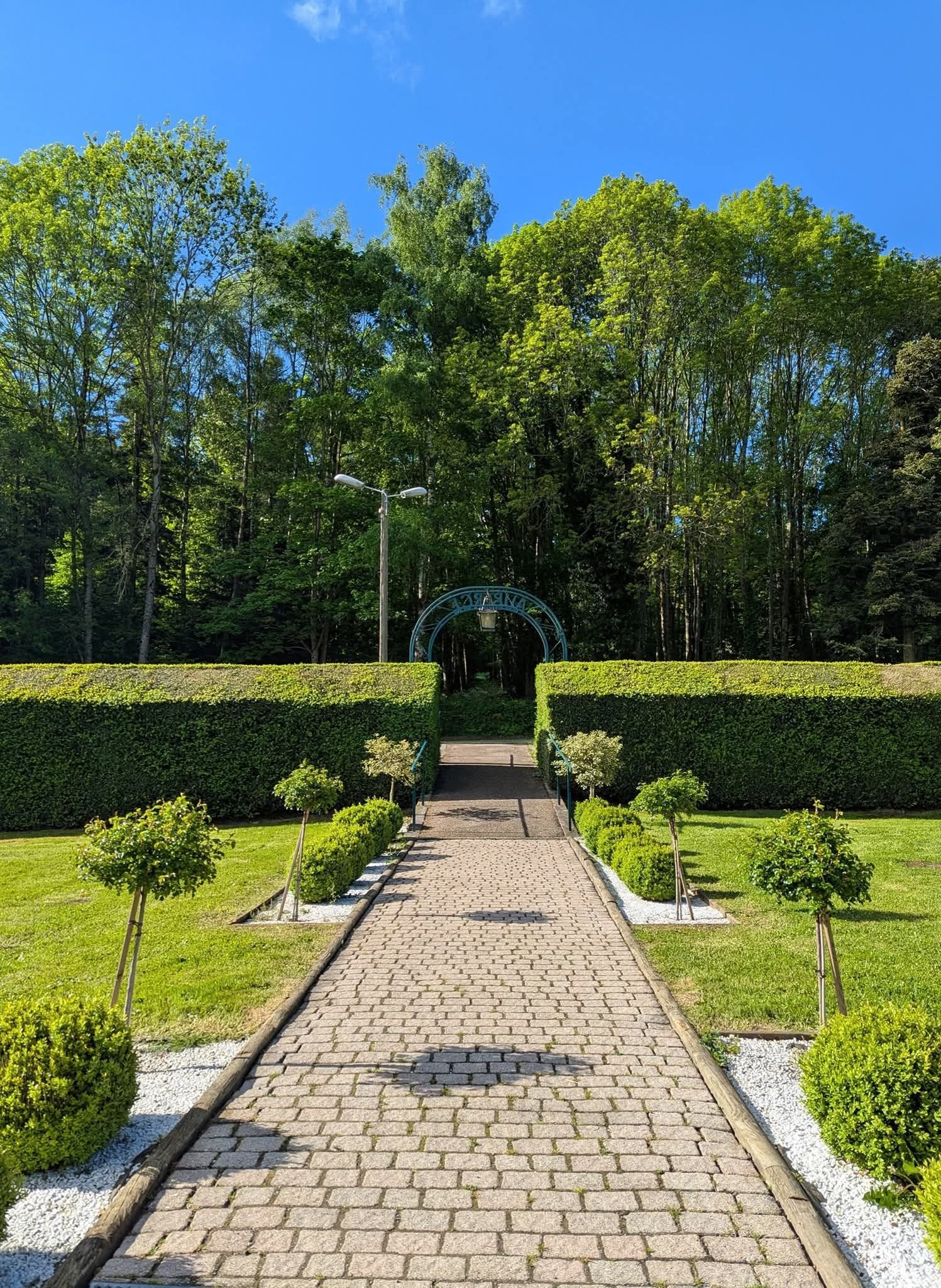 Un chemin de pierre traverse un jardin soigné et mène à une arche métallique encadrée de haies denses, sous un ciel d'un bleu éclatant.