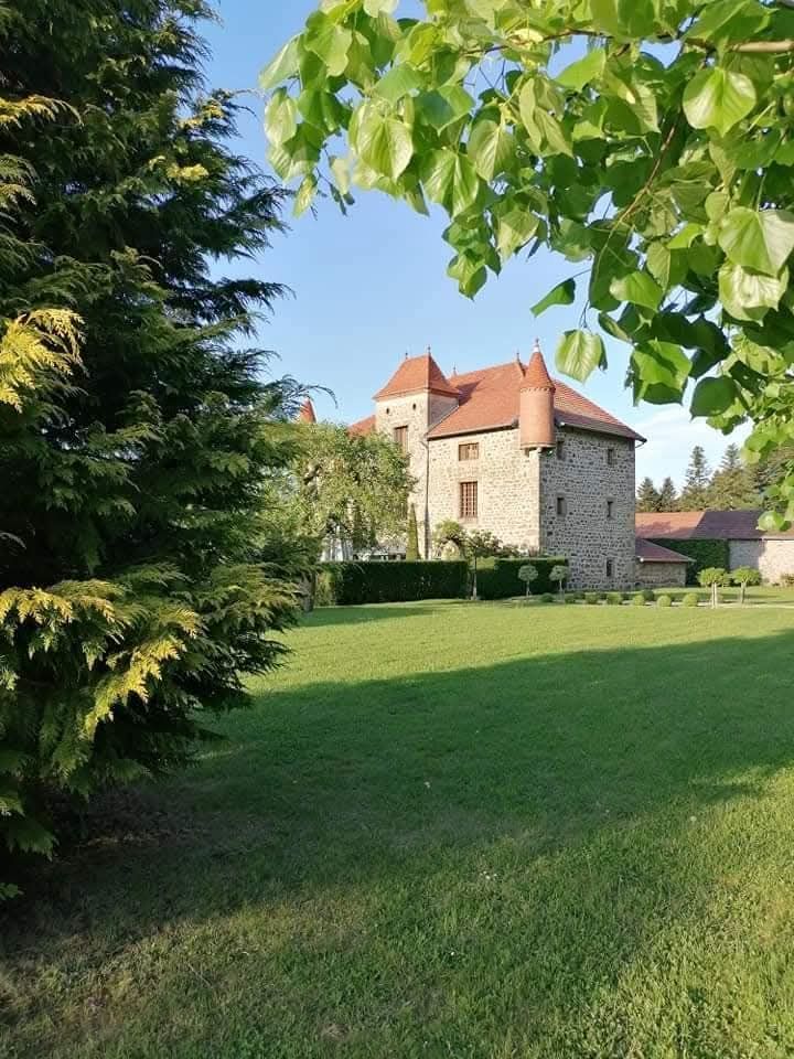 Un grand château en pierre au toit de tuiles rouges se dresse derrière une pelouse verdoyante et des arbres par une journée ensoleillée.