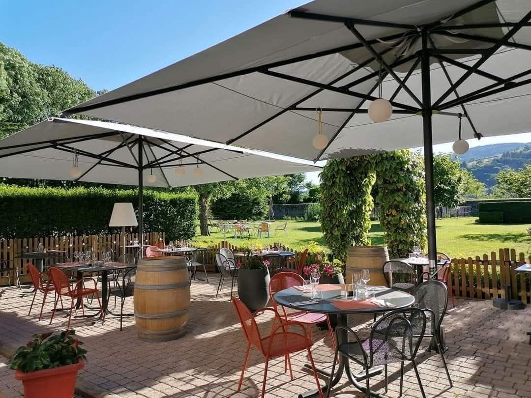 Une terrasse extérieure avec des tables en métal, des chaises orange et des tables en forme de tonneau, ombragée par de grands parasols, dans un cadre de jardin.