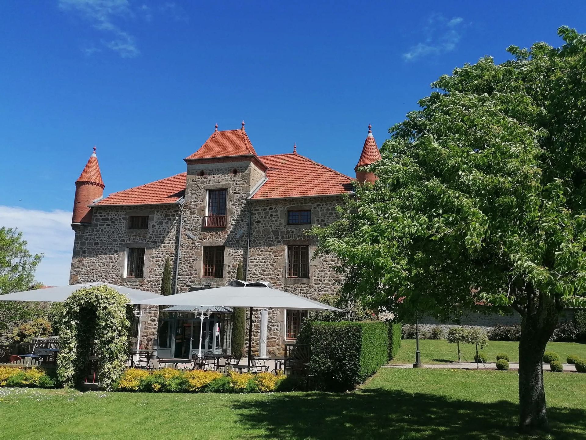 Un château en pierre au toit de tuiles rouges et doté de deux tours d'angle, situé dans un jardin ensoleillé avec des parasols sur la pelouse.