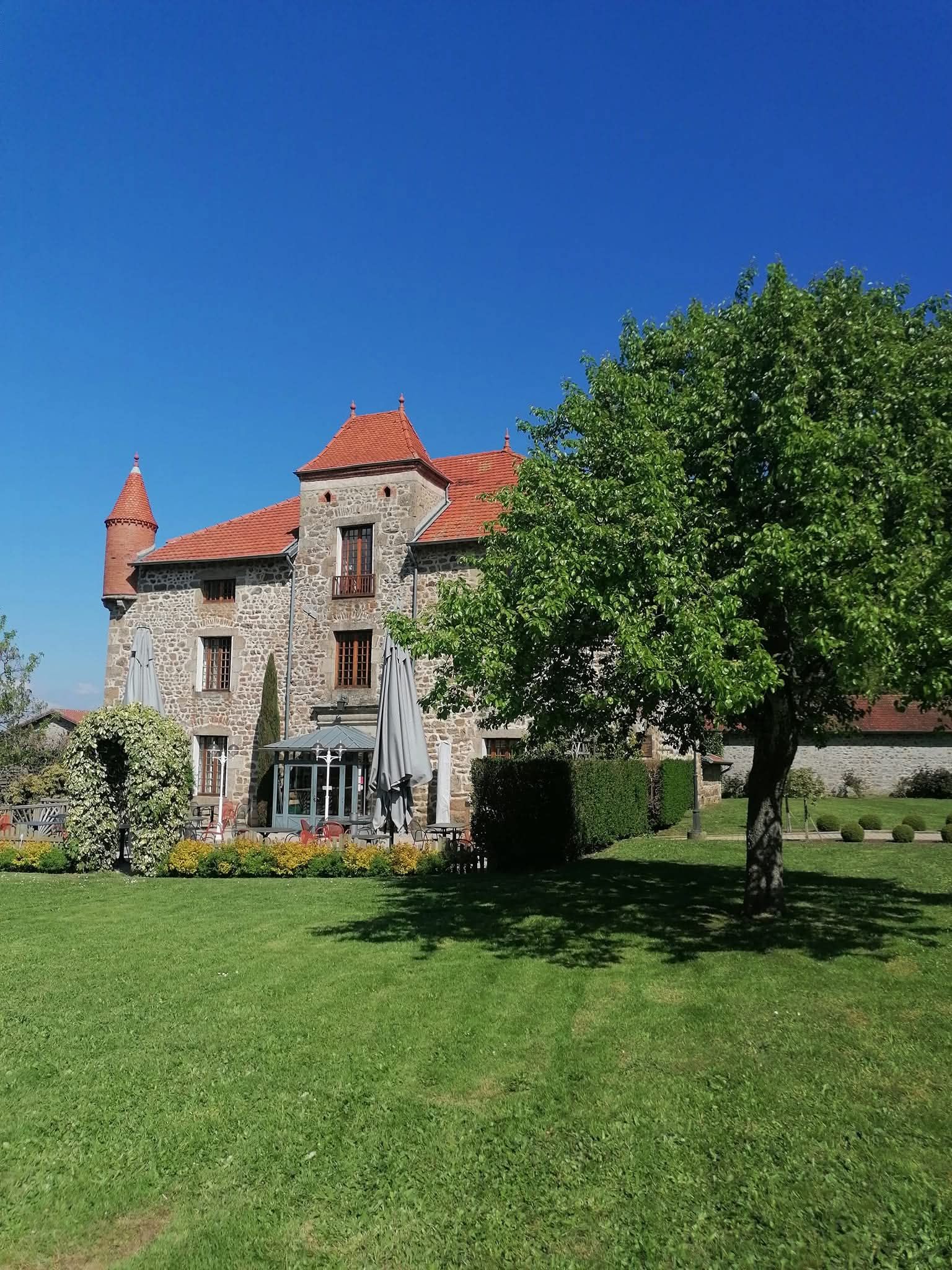 Un manoir en pierre, au toit de tuiles rouges et surmonté d'une tourelle, se dresse derrière une pelouse verdoyante et un grand arbre, sous un ciel d'un bleu éclatant.