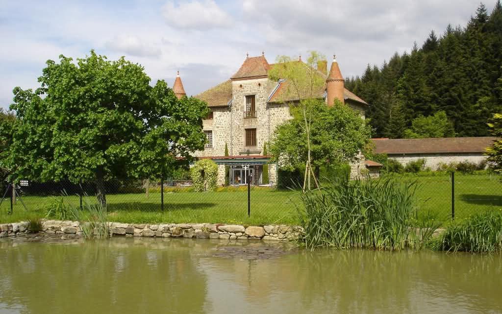 Un château de pierre aux tours pointues se dresse derrière une pelouse verdoyante et un étang, sous un ciel partiellement nuageux.