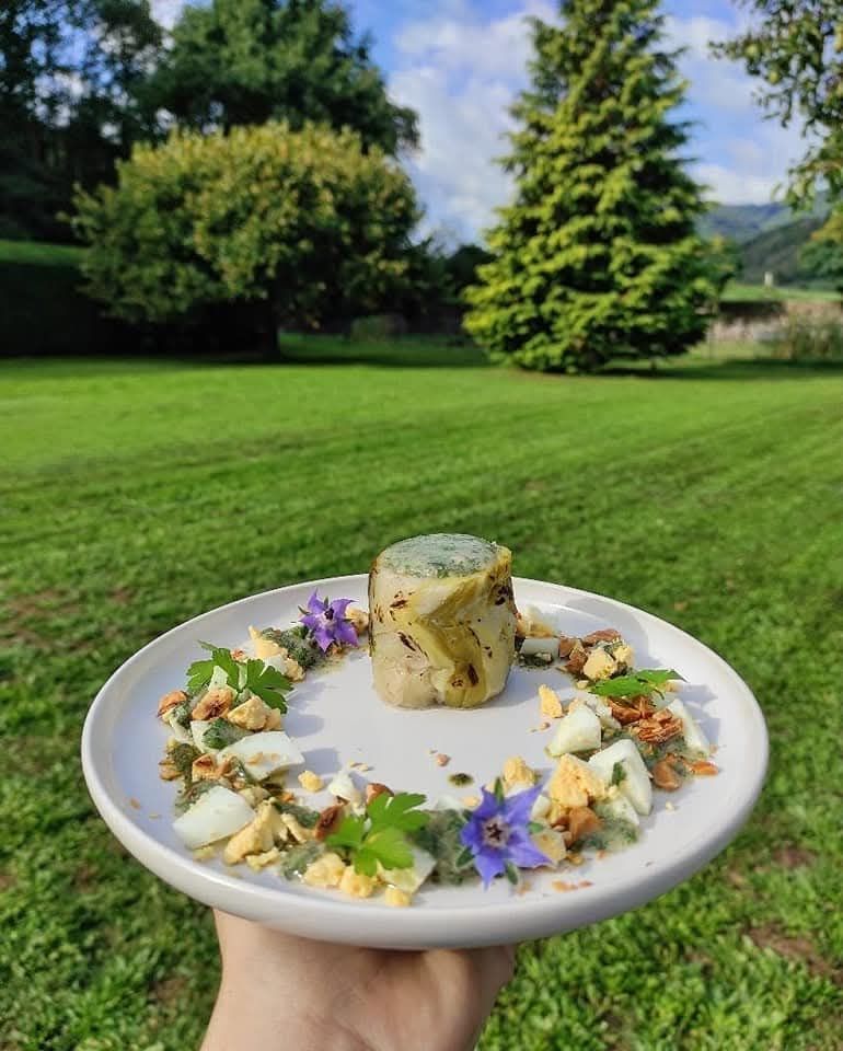 Une main tient une assiette sur laquelle est disposée une terrine cylindrique verte garnie de fleurs comestibles et de noix, dans un champ ensoleillé.