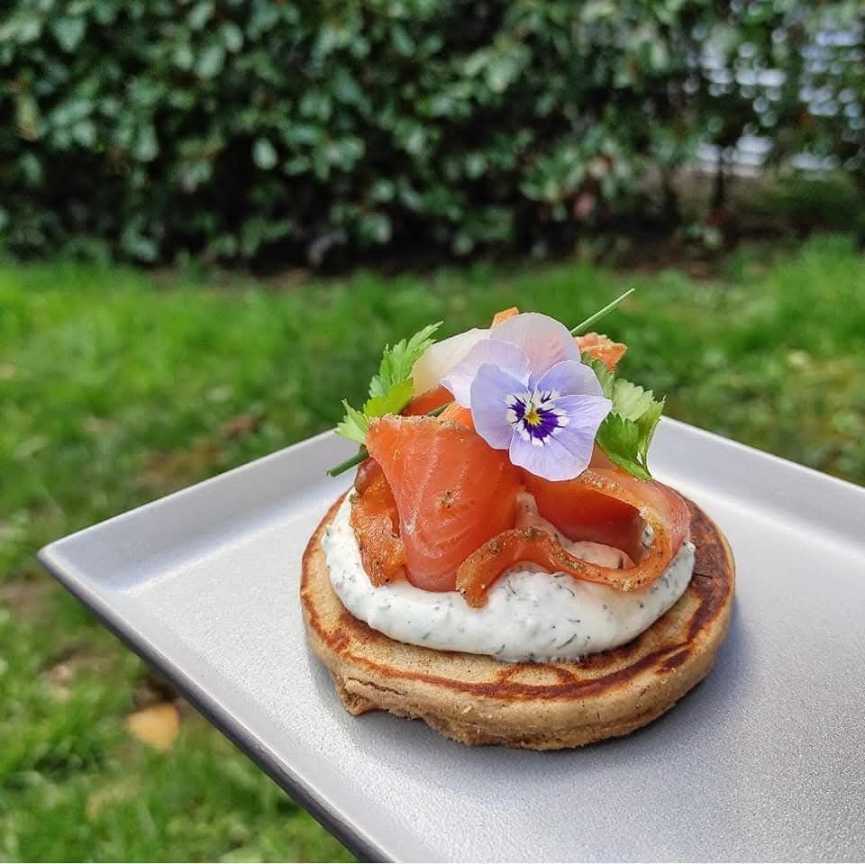 Un blini au saumon fumé, nappé de crème aux herbes, garni d'une garniture et d'une petite fleur violette, servi sur une assiette en extérieur.