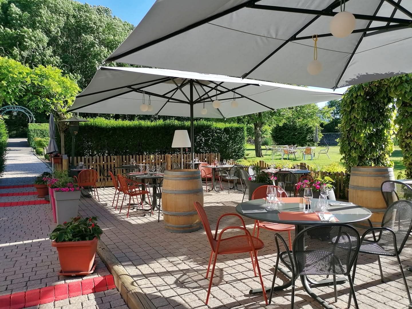 Terrasse extérieure avec tables, chaises orange et parasols blancs, dans un cadre ensoleillé évoquant un jardin.
