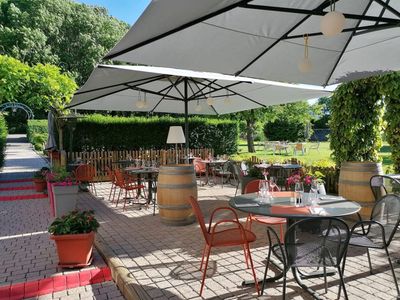 Terrasse extérieure de restaurant avec tables, chaises et grands parasols par une journée ensoleillée, avec des arbres verts en arrière-plan.