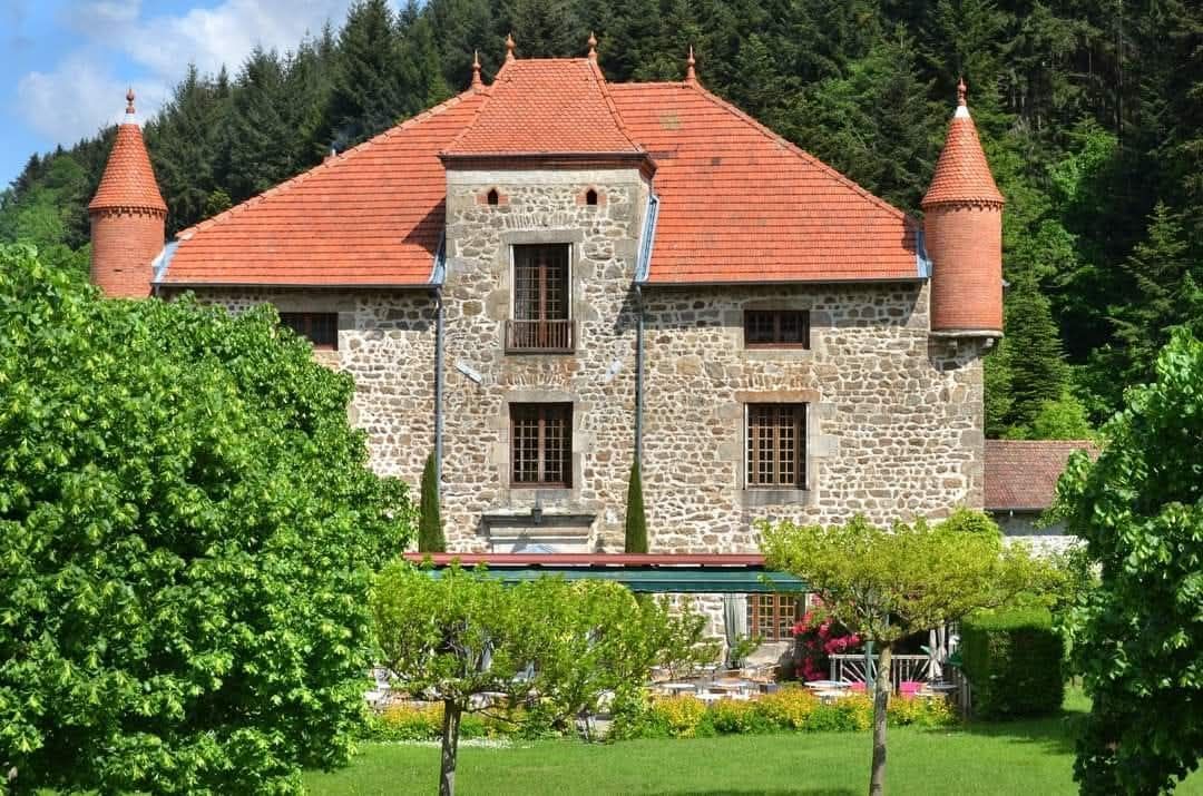 Un château en pierre au toit de tuiles rouges et doté de deux tourelles d'angle, entouré d'une végétation luxuriante et d'un jardin.
