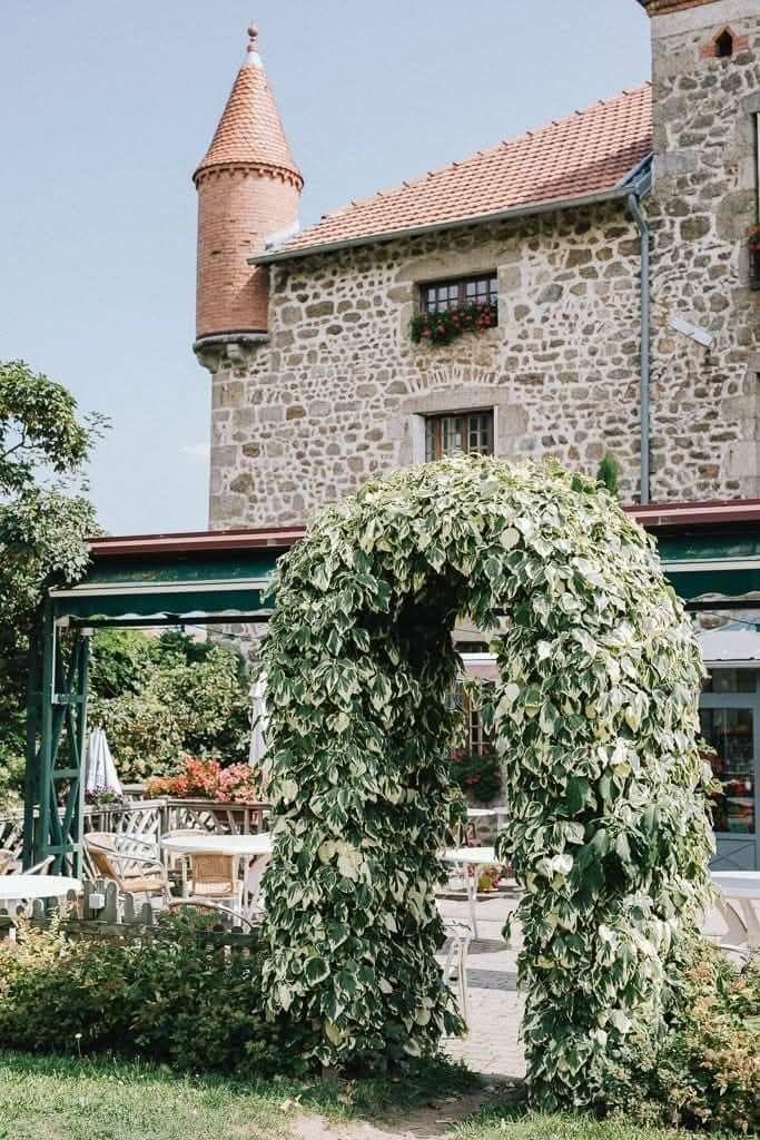 Une treille voûtée recouverte de vigne se dresse devant un bâtiment en pierre doté d'une tourelle proéminente, sous un ciel d'un bleu limpide.