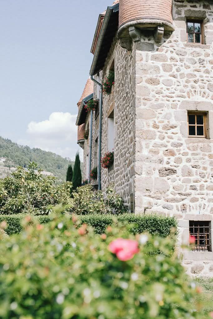 Un bâtiment en pierre, orné de tourelles et de petites fenêtres, se dresse à côté d'un jardin verdoyant luxuriant où une unique fleur rose s'épanouit au premier plan.