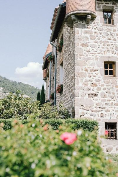 Un château aux murs de pierre, surmonté d'une tourelle, se dresse derrière une haie verte et une rose rose, avec en arrière-plan des collines lointaines.