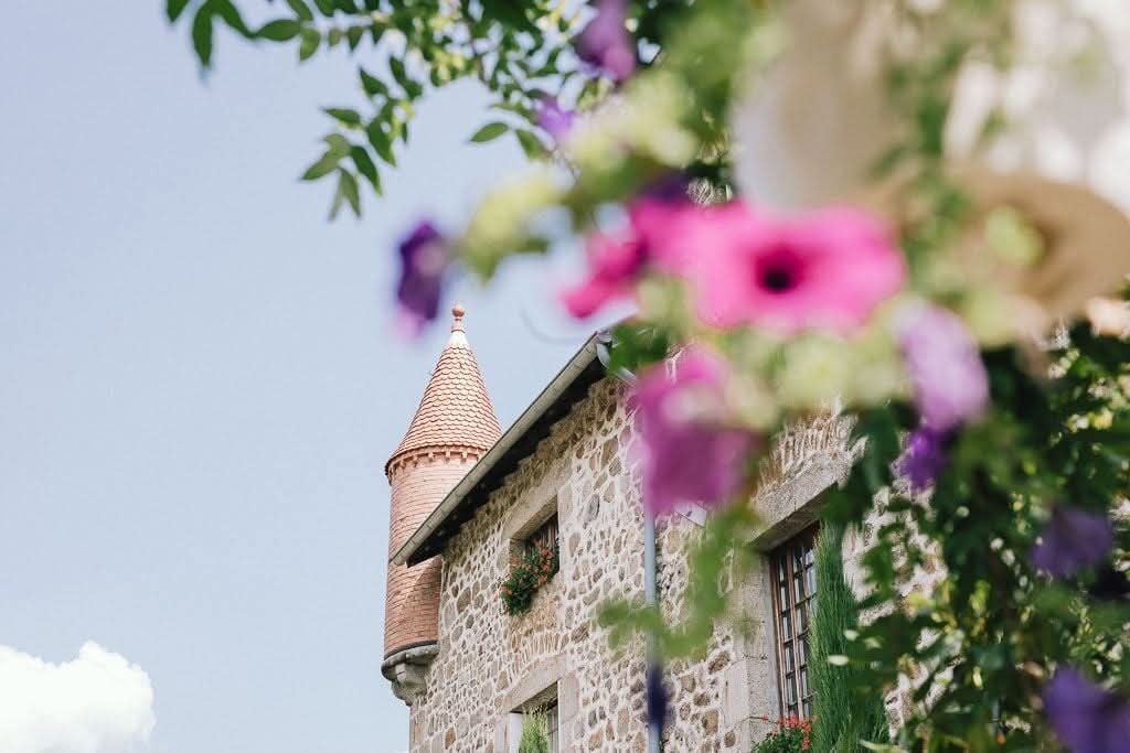 Un bâtiment en pierre avec une tourelle pointue, partiellement masqué par des fleurs suspendues d'un rose et d'un violet éclatants.