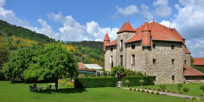 Un bâtiment en pierre, doté de deux tourelles d'angle et d'un toit de tuiles rouges, se dresse derrière une pelouse, des arbres et une haie basse.