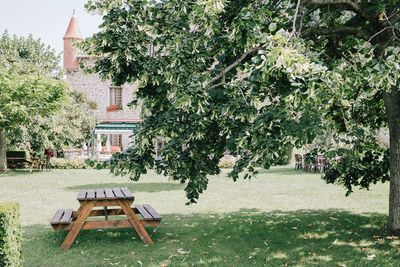 Une table de pique-nique en bois est installée sur une pelouse herbeuse, à l'ombre d'un grand arbre, devant un bâtiment en pierre surmonté d'une tourelle.