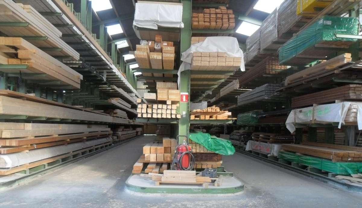 Intérieur d'un entrepôt avec de hautes étagères métalliques remplies de bois et de palettes emballées dans du papier vert, vu depuis une allée centrale.
