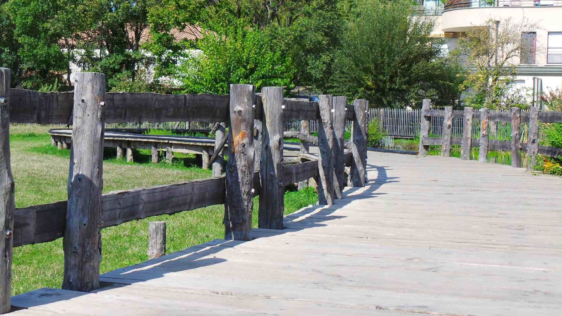 Une clôture en bois borde une promenade ensoleillée, à côté d'arbres verdoyants et d'un parc gazonné.