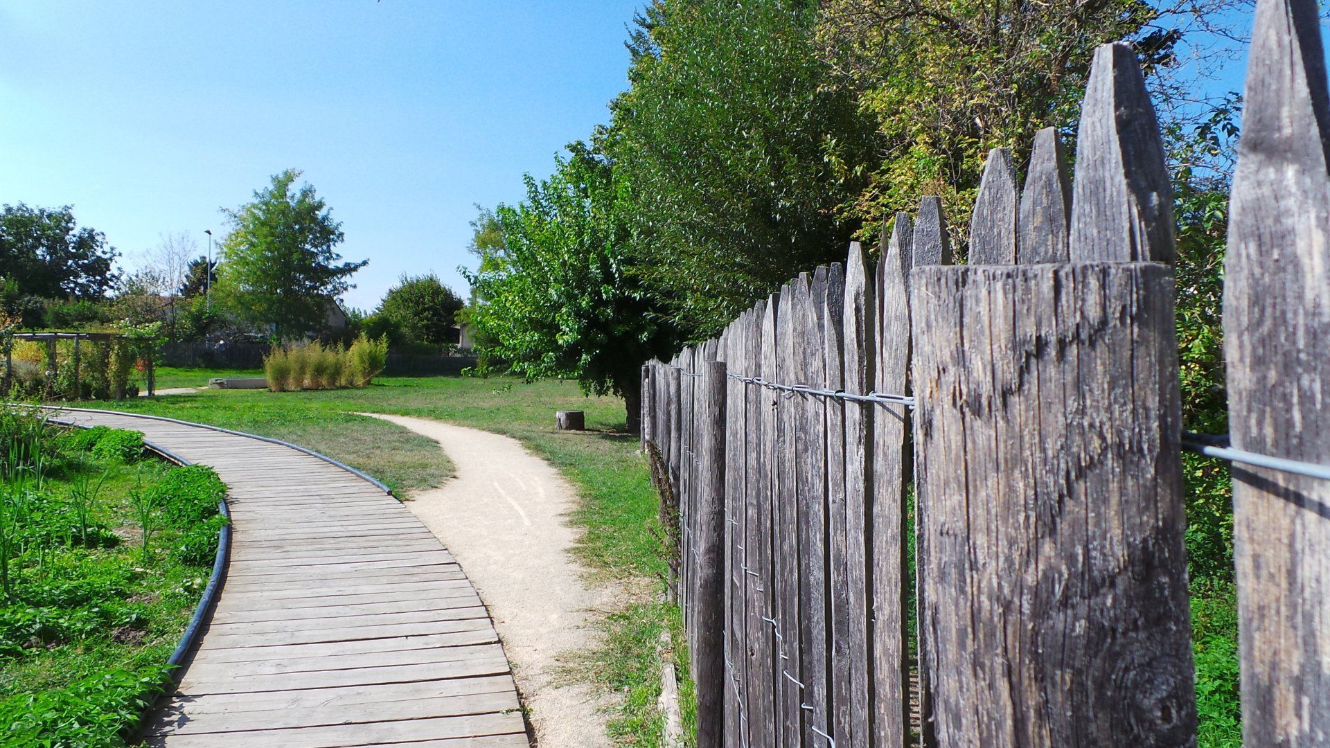 Chemin de pierres sinueux bordé d'herbe verte et d'arbres, avec une clôture en bois patinée par le temps sur la droite.