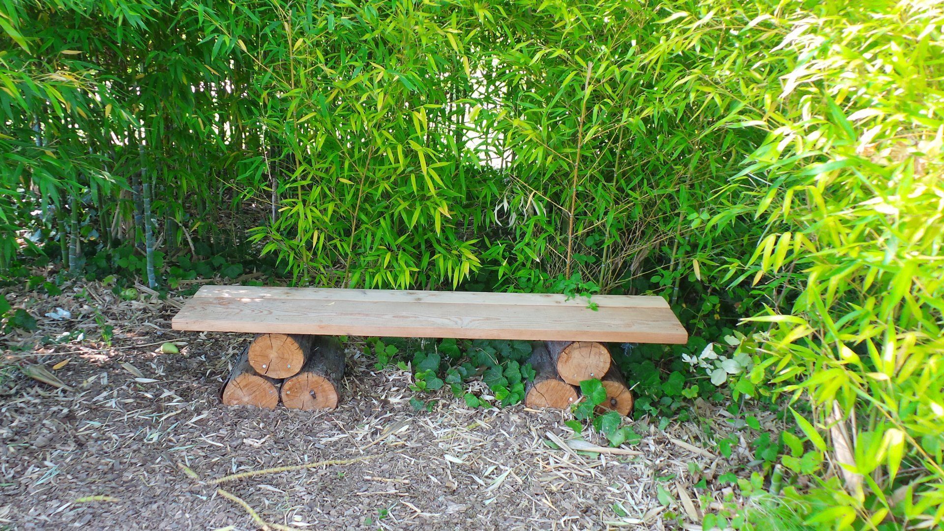 Banc en bois dans un jardin arboré, soutenu par des pierres empilées sur un chemin de gravier.