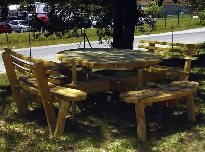 Table de pique-nique en bois avec bancs sous des arbres dans un parc, avec une route et des voitures en arrière-plan.