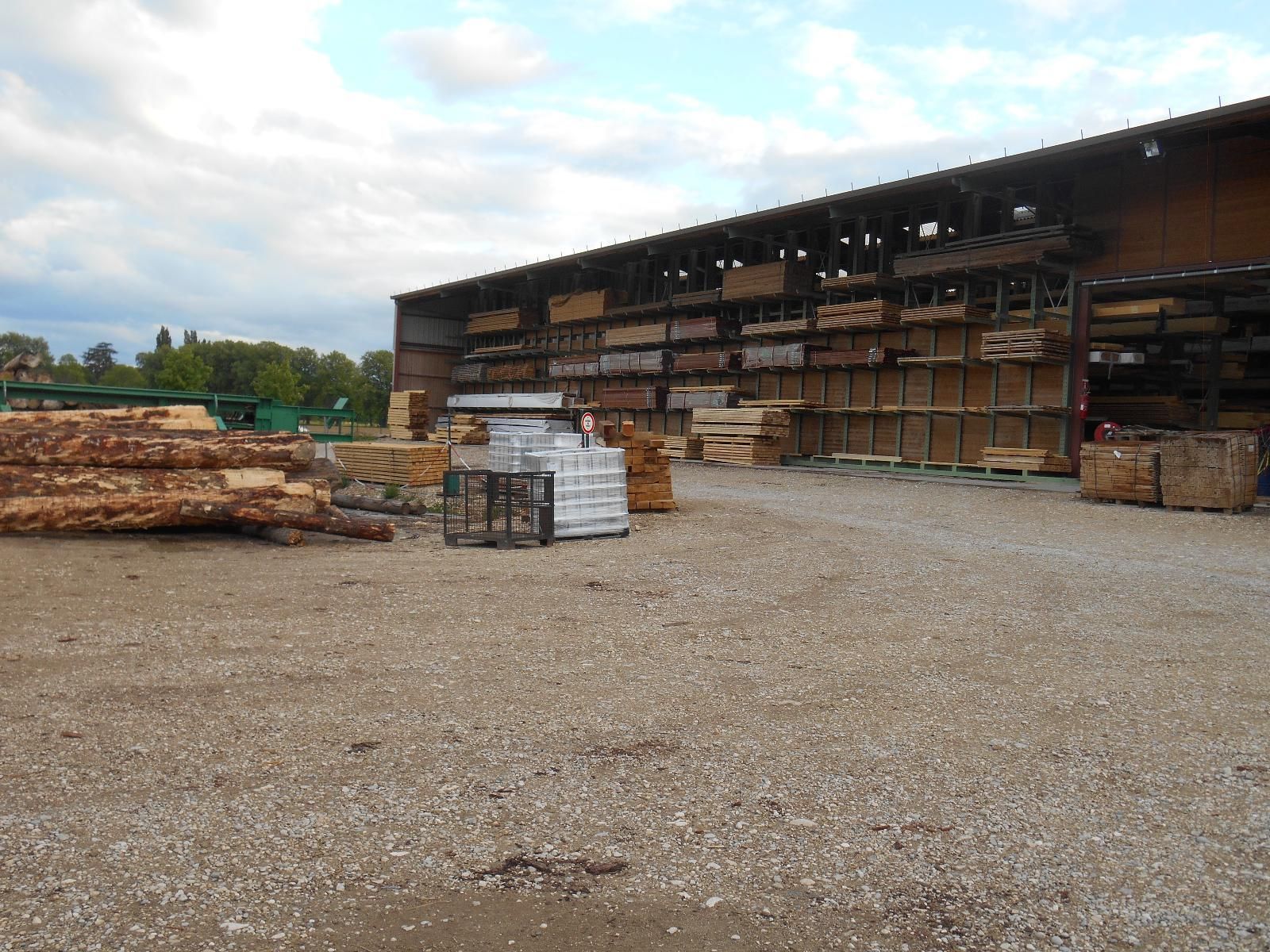 Terrain industriel avec du bois d'œuvre et des palettes empilés sous un hangar couvert, sur un sol gravillonné.
