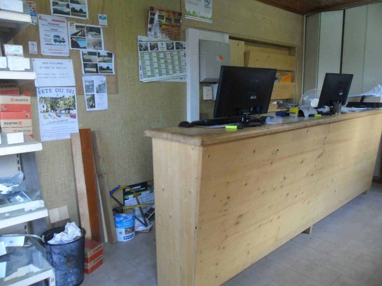 Petit espace d'accueil avec un comptoir en bois, des écrans d'ordinateur et des affiches au mur.
