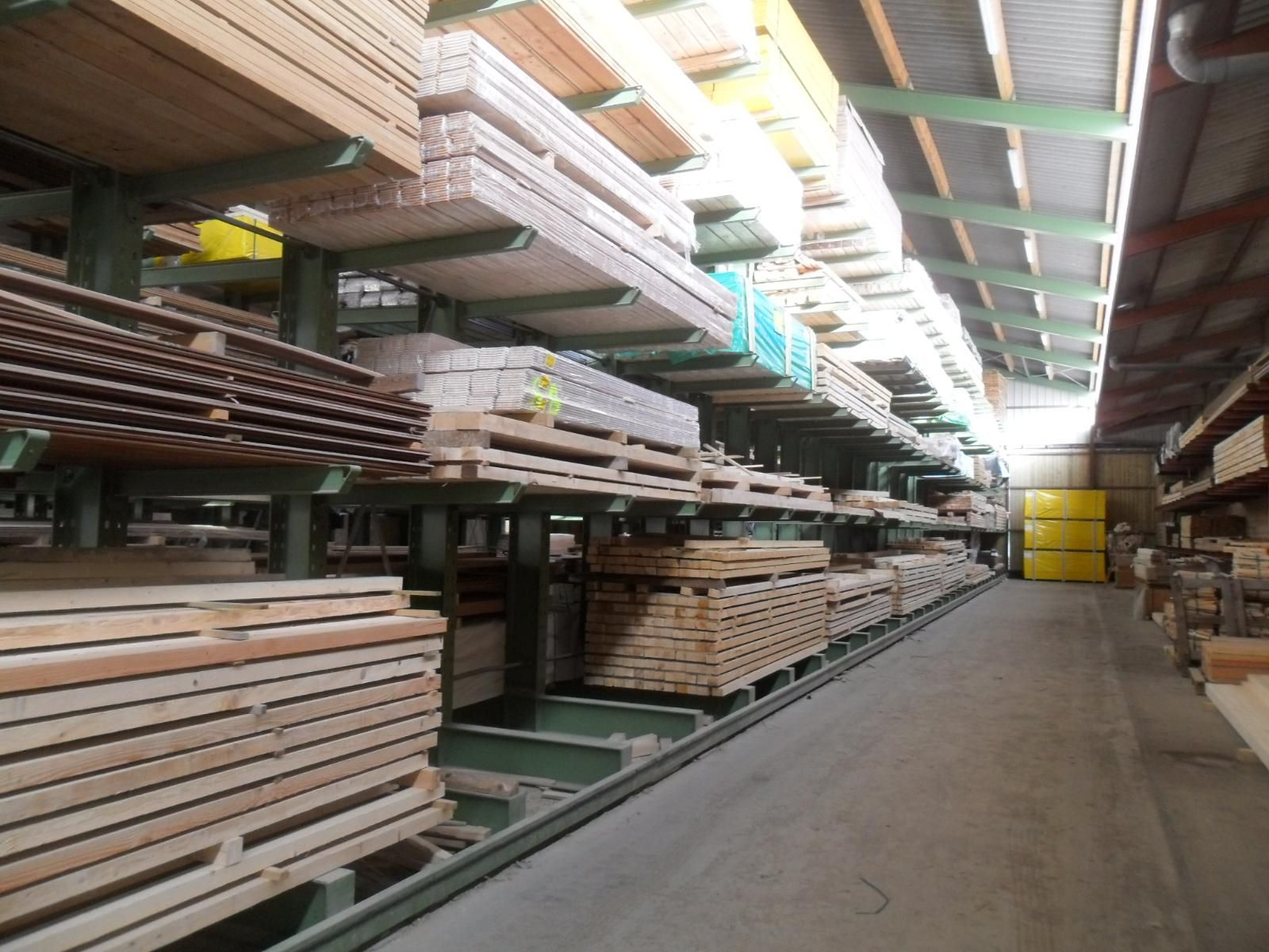 Allée d'un entrepôt de bois avec des planches empilées sur des étagères et des espaces de stockage en hauteur, éclairée par des puits de lumière.