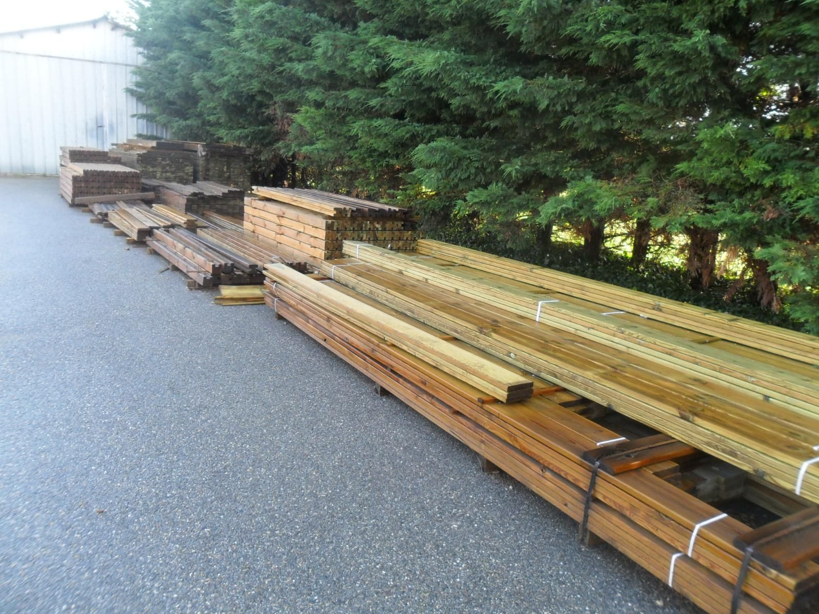Des piles de bois et de poutres métalliques sont entreposées le long d'un chemin de gravier, à côté d'arbres et d'un bâtiment blanc.