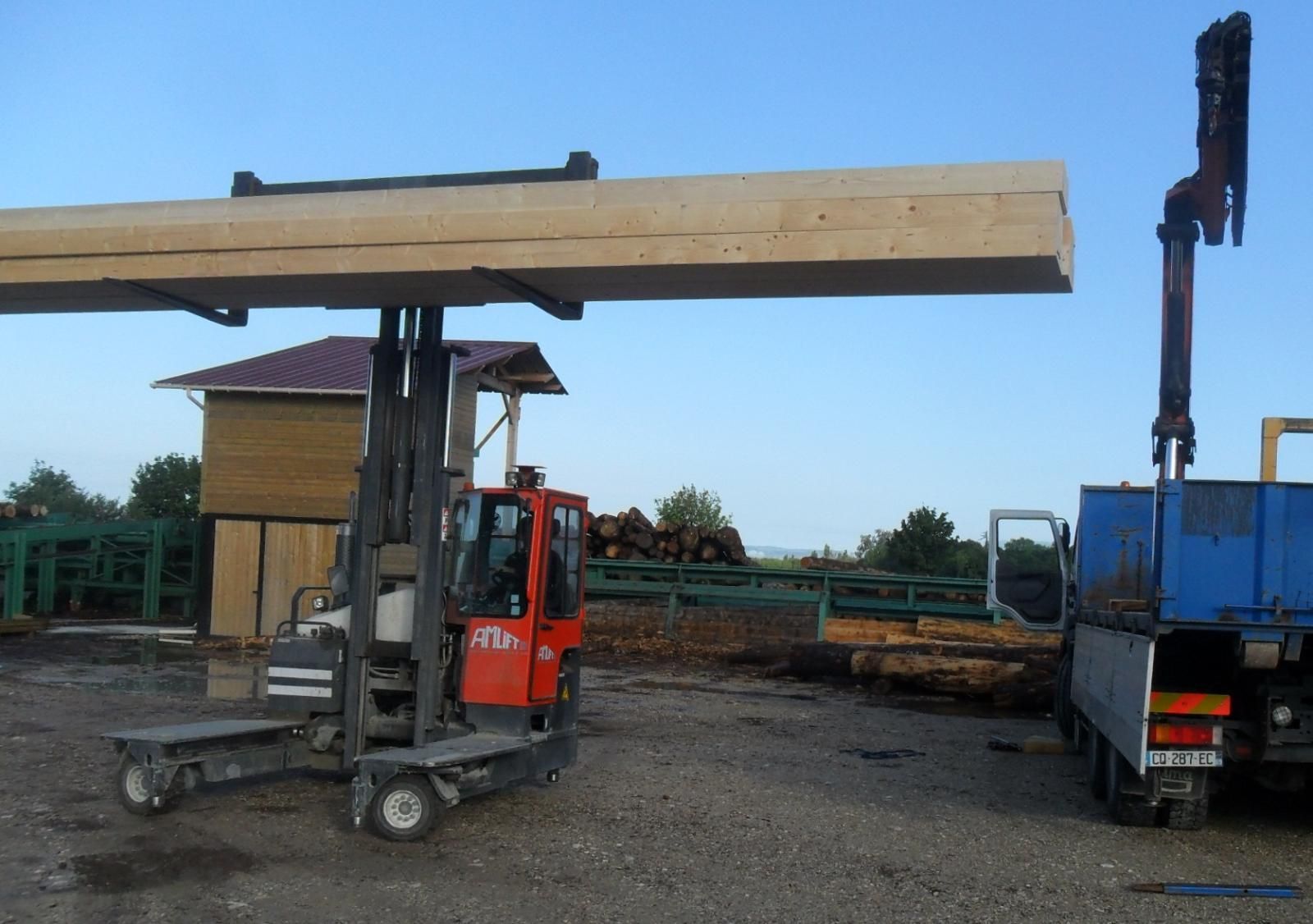 Un chariot élévateur soulève une longue poutre en bois à côté d'un camion bleu dans une zone industrielle.