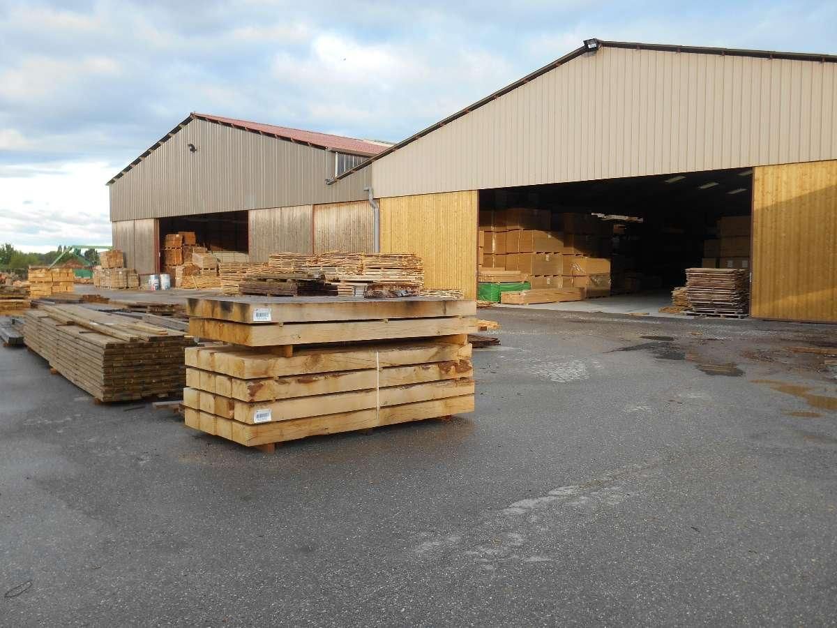 Parc à bois avec des fagots de bois empilés à l'extérieur de grands hangars de stockage ouverts sur les côtés.