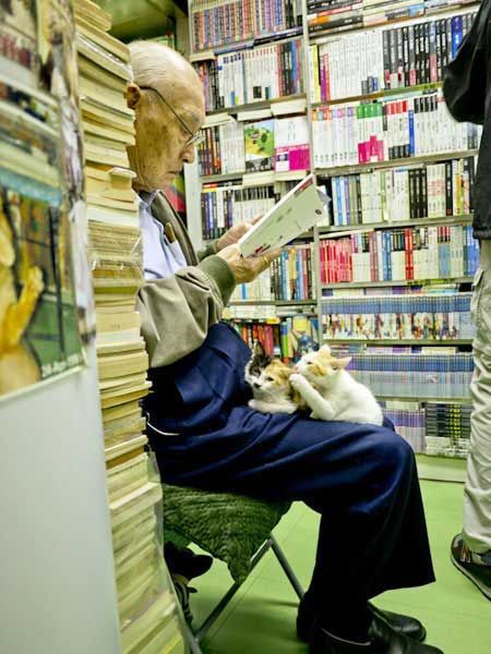 Un homme lit un livre dans une allée étroite d'une librairie, bordée d'étagères de livres des deux côtés.