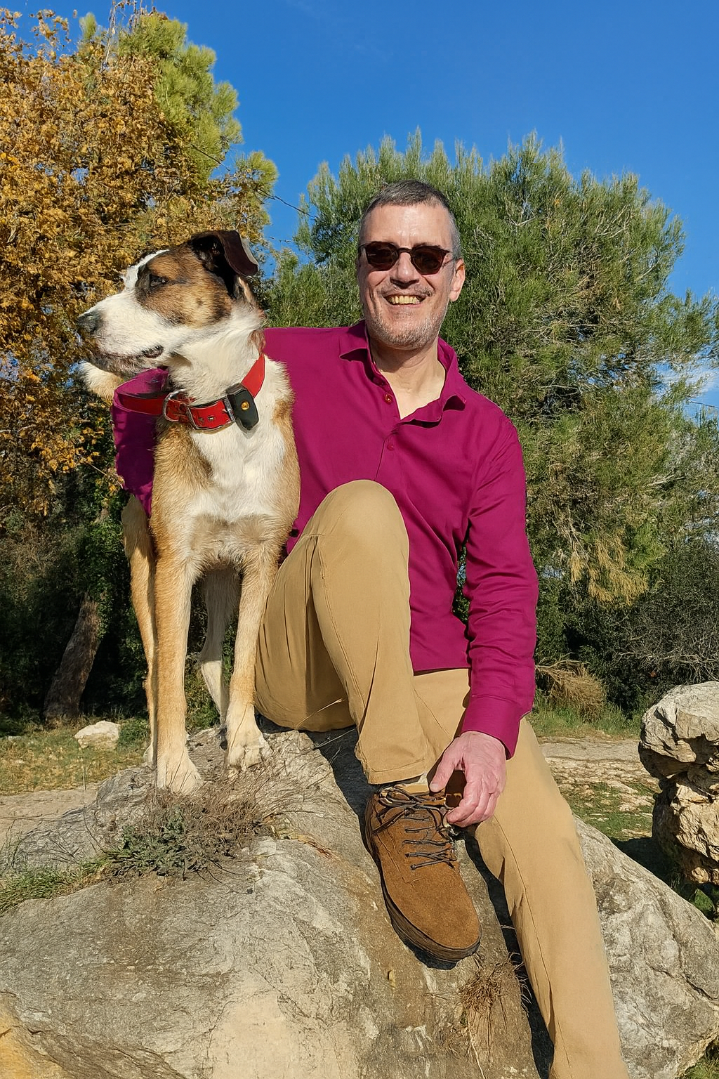 Un homme en chemise rose et pantalon beige est assis à côté d'un chien sur un rocher, tous deux souriant en plein air.